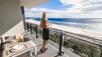A balcony with a view of the beach, a table set with plates, a bottle of champagne, and a glass.