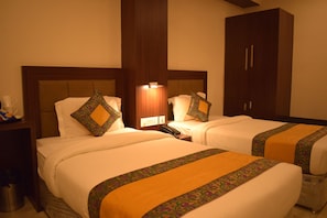 A hotel room with two beds, a wooden wardrobe, a bedside table with a lamp, and a trash bin. - Hari Vilaas (Varanasi)