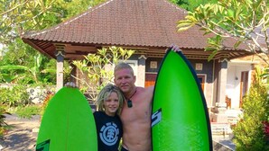 Un homme et un enfant, chacun tenant une planche de surf, se tiennent devant une maison avec un toit en tuiles et une véranda.