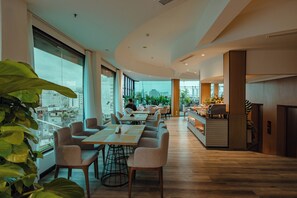 Un espace de restauration moderne avec de longues tables et des chaises, de grandes fenêtres offrant une vue sur la ville et un comptoir de cuisine où sont disposés des aliments.