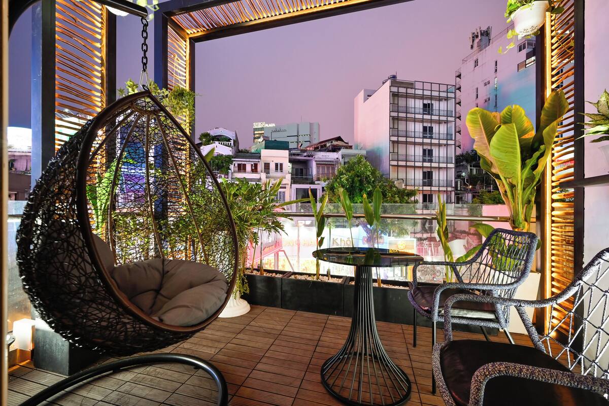 A balcony with a hanging chair, a small table, and chairs, overlooking a cityscape with buildings and a view of the sky.