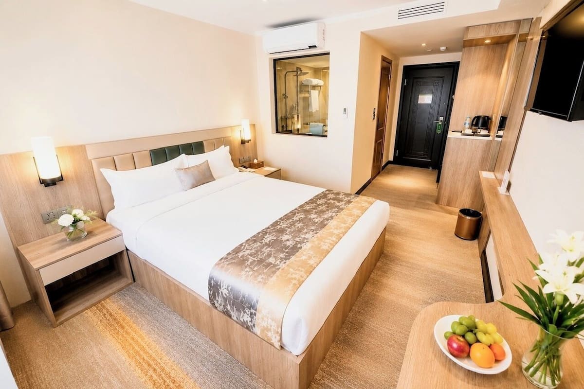A hotel room with a bed, bedside table, nightstand, and a small table with fruit and flowers.