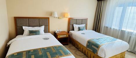 A hotel room with two beds, a nightstand, a lamp, and a window with curtains.