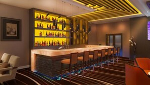 Zona de bar con una barra larga y varios taburetes. Una pared exhibe botellas en estantes. La habitación tiene una alfombra a rayas y un diseño de techo moderno.