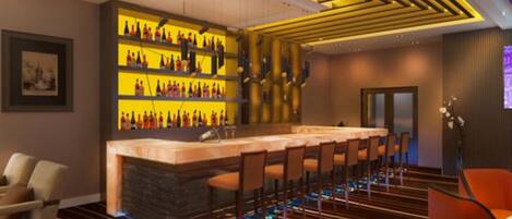 A bar area with a long counter and multiple bar stools. There is a wall with shelves displaying bottles. The room has a striped carpet and a modern ceiling design.