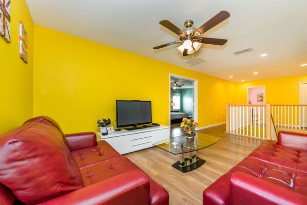 A living room with a red leather couch, a glass coffee table, a flat-screen TV, and a ceiling fan.