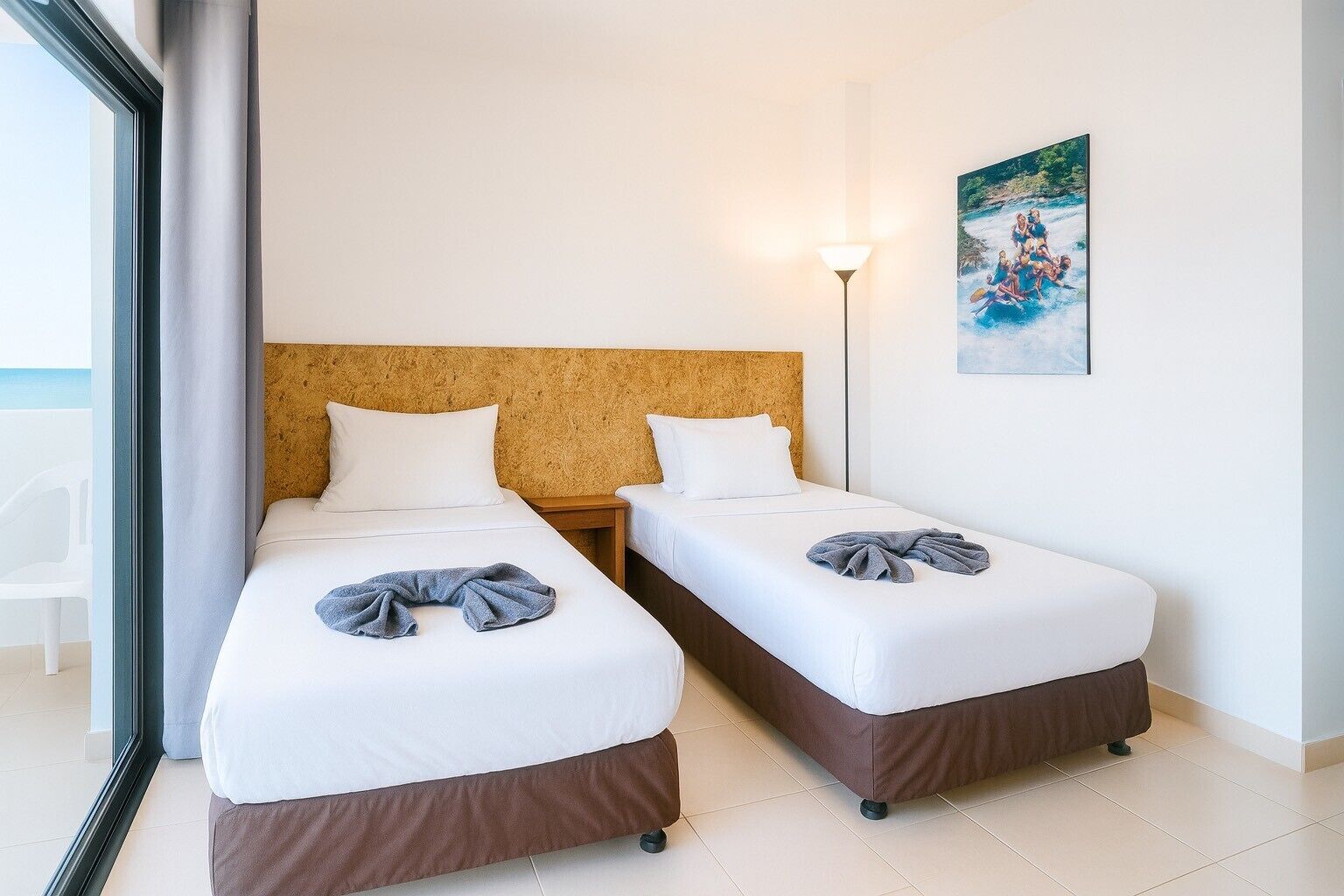 A hotel room with two single beds, a large window, a painting on the wall, and a lamp.