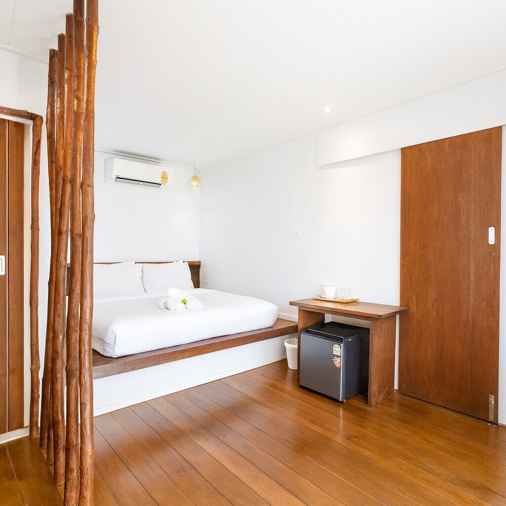 A modern hotel room with a wooden floor, a bed with white linens, a small desk with a computer, and a wardrobe.