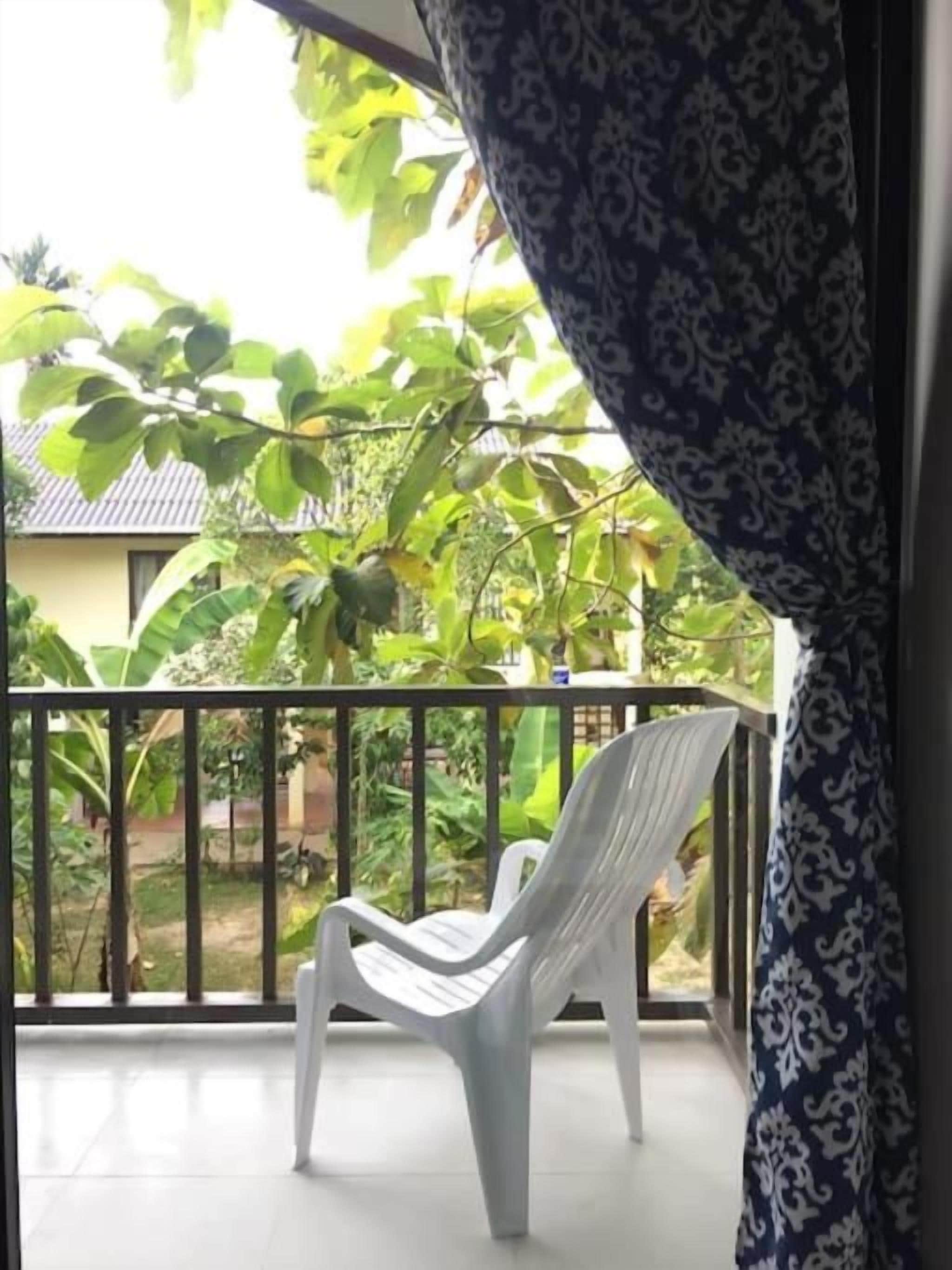 Un balcón con una silla de plástico blanca, una cortina estampada y vistas a una casa y follaje.