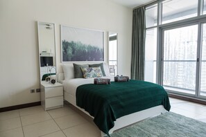 A modern hotel room with a large window, a bed with a green bedspread, a bedside table with a lamp, and a large framed picture on the wall.