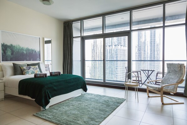A modern bedroom with a large window, a bed with a green blanket, a chair, and a small table.