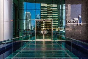 A modern hotel entrance with a glass staircase, a prominent 'indigo tower' sign, and a view of surrounding buildings.