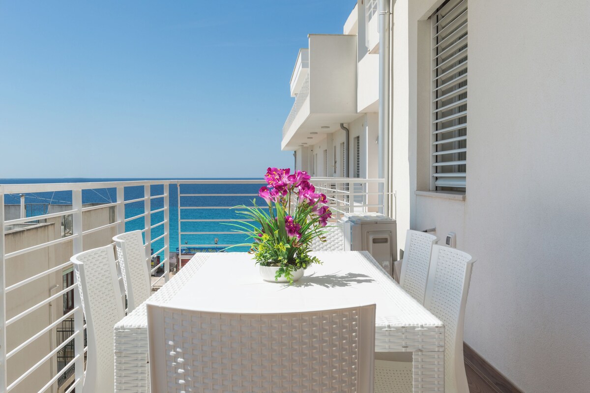 A balcony with a white table and chairs, a potted plant with flowers, and a view of the sea.