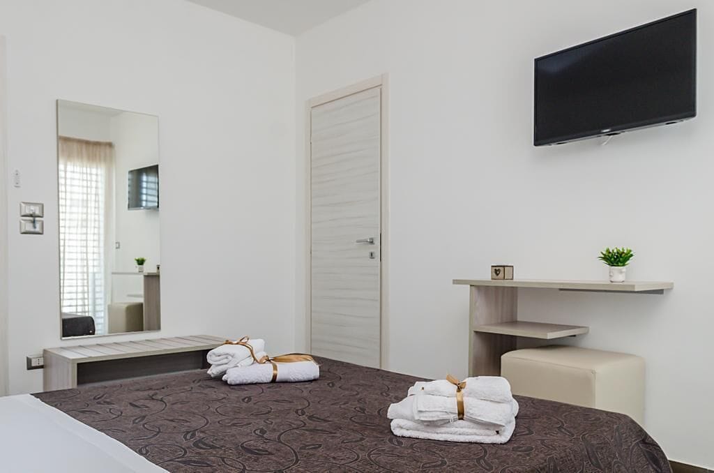 Ein Hotelzimmer mit Bett, Wandfernseher, Spiegel, Nachttisch mit Pflanze und Tür.