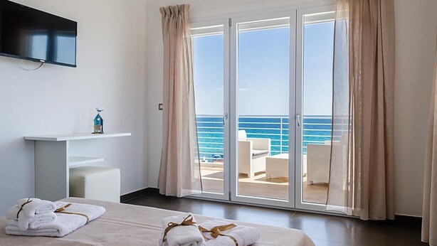 Una habitación de hotel con cama, televisor montado en la pared, balcón con vista al mar y una pequeña estantería con una botella.