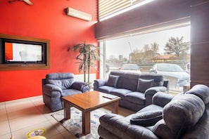 Un salon avec un mur rouge, un téléviseur, une grande fenêtre donnant sur des voitures garées et une plante en pot.