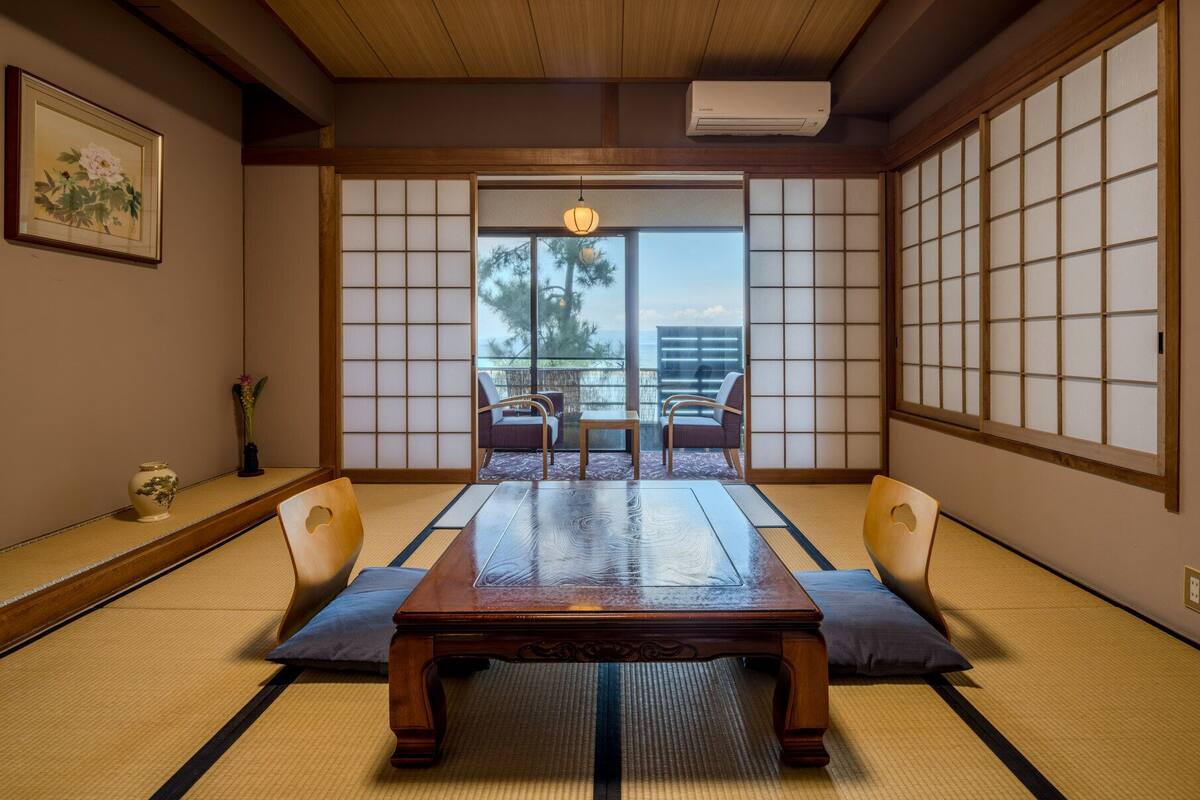A traditional Japanese room with tatami flooring, a low table, two chairs, a shoji screen, and a view of the outdoors.