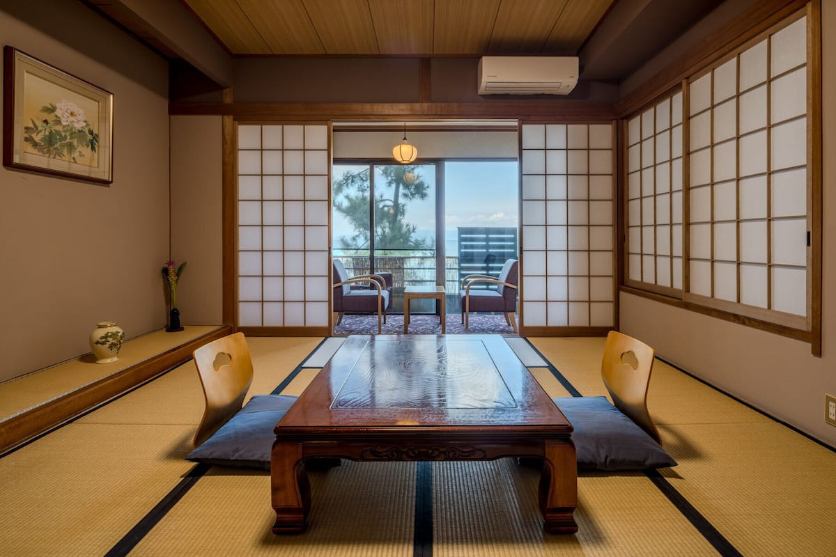 A traditional Japanese room with tatami flooring, a low table, two chairs, a shoji screen, and a view of the outdoors.