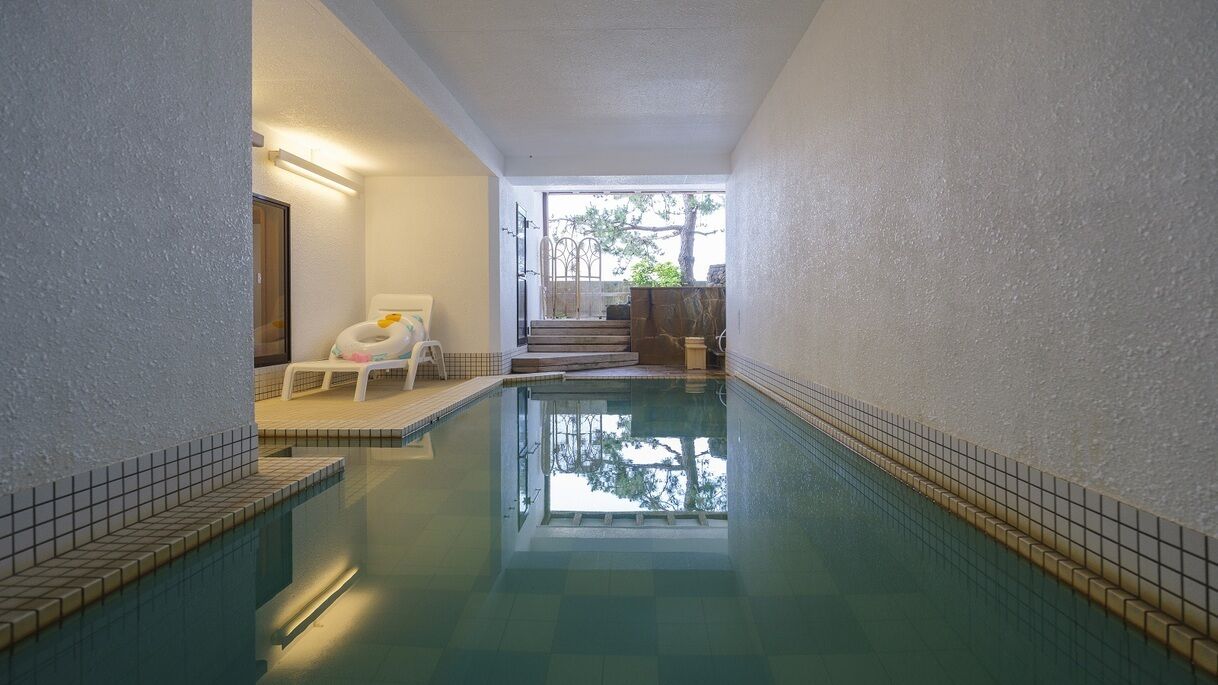 En indendørs swimmingpool med flisekant, en liggestol og en trappe der fører til et udendørs område.