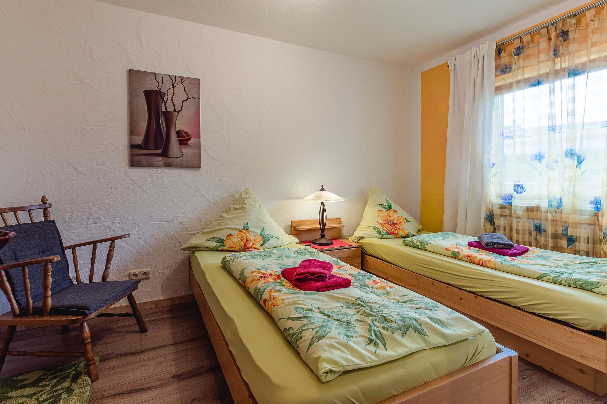 A room with two single beds, a wooden chair, a table lamp, and a painting on the wall.