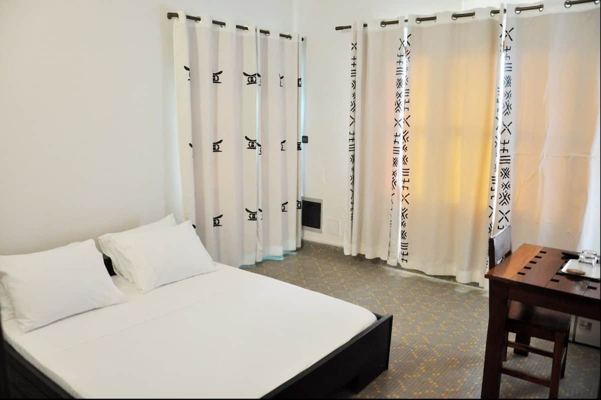A hotel room with a bed, white curtains with black patterns, a wooden table, and a carpeted floor.
