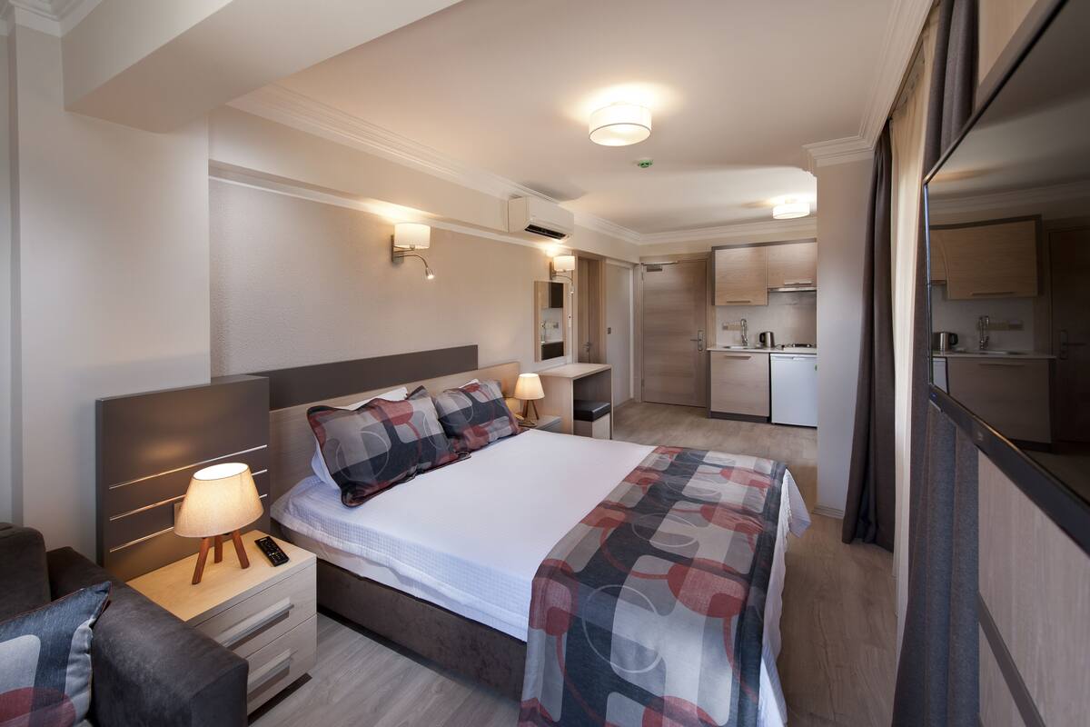 A modern hotel room with a bed, bedside table, lamp, and a view of the kitchen.