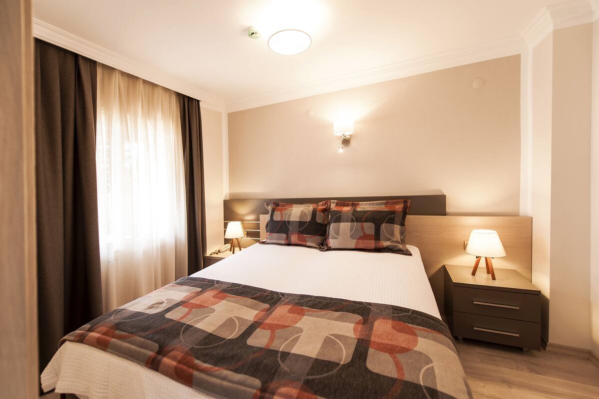 A hotel room with a large bed, two bedside tables with lamps, a headboard, and a window with curtains.