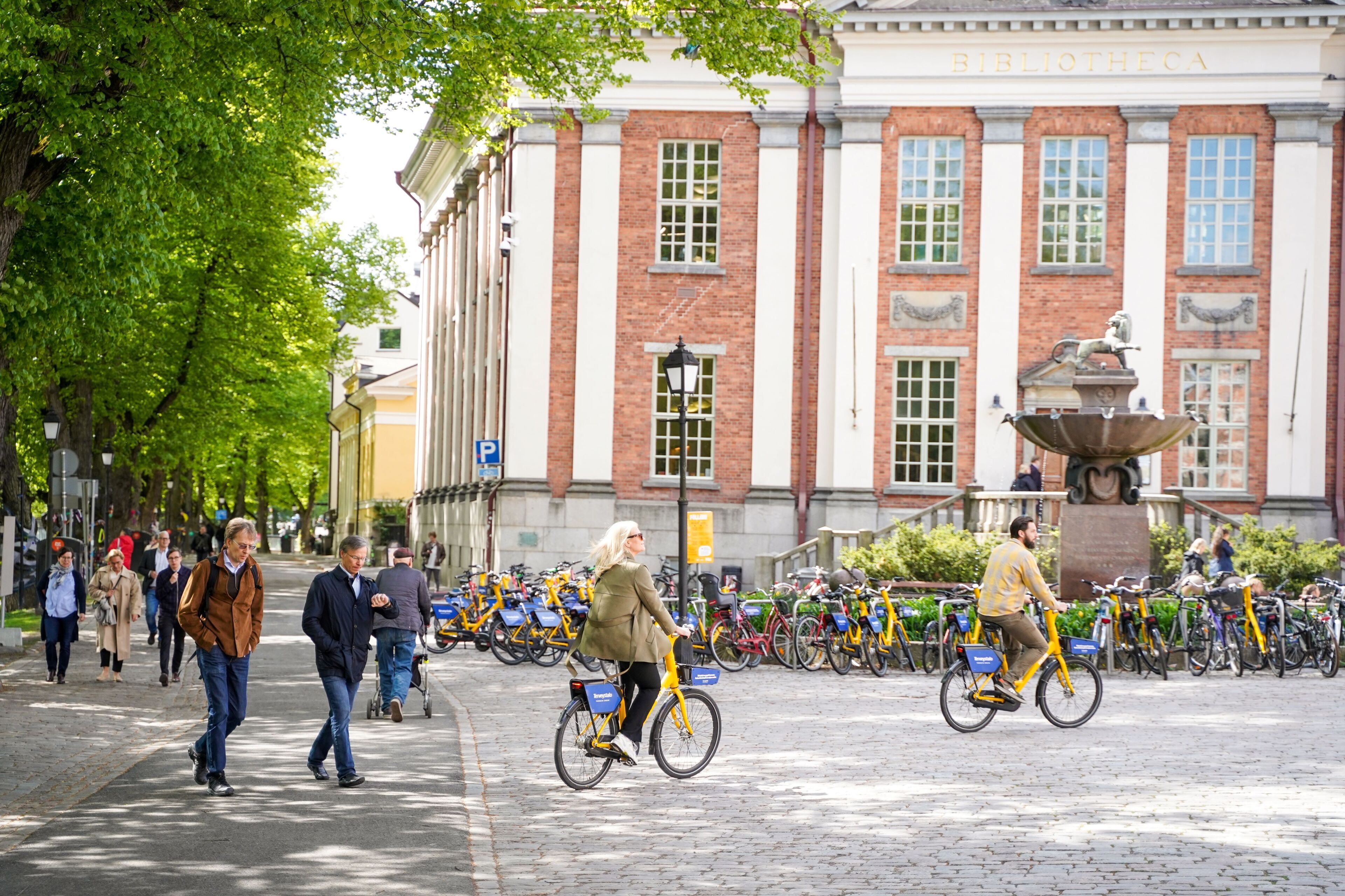 A street scene with pedestrians and cyclists, a building with a sign reading "BIBLIOTHECA", and a fountain in the background.