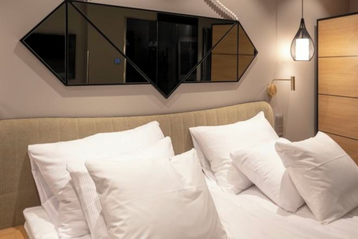 A hotel room with a bed, white pillows, a wooden headboard, a wall-mounted mirror, and a pendant light.