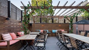 Une terrasse extérieure avec des tables et des bancs en bois, une pergola illuminée de guirlandes lumineuses, et une vue sur des arbres et un bâtiment en arrière-plan.