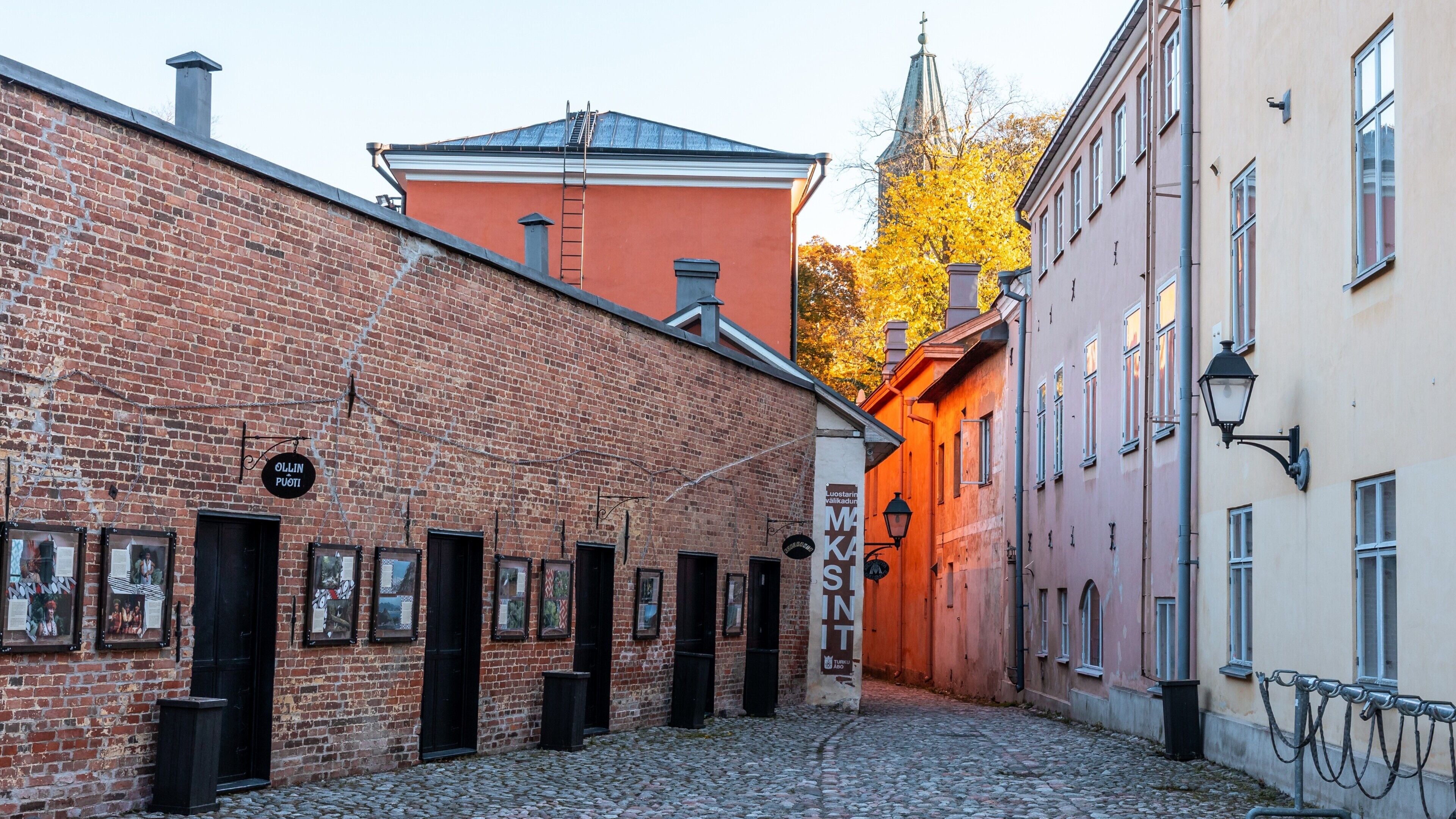 A cobblestone street with a brick building, a red building with a tower, and a yellow building with a sign.