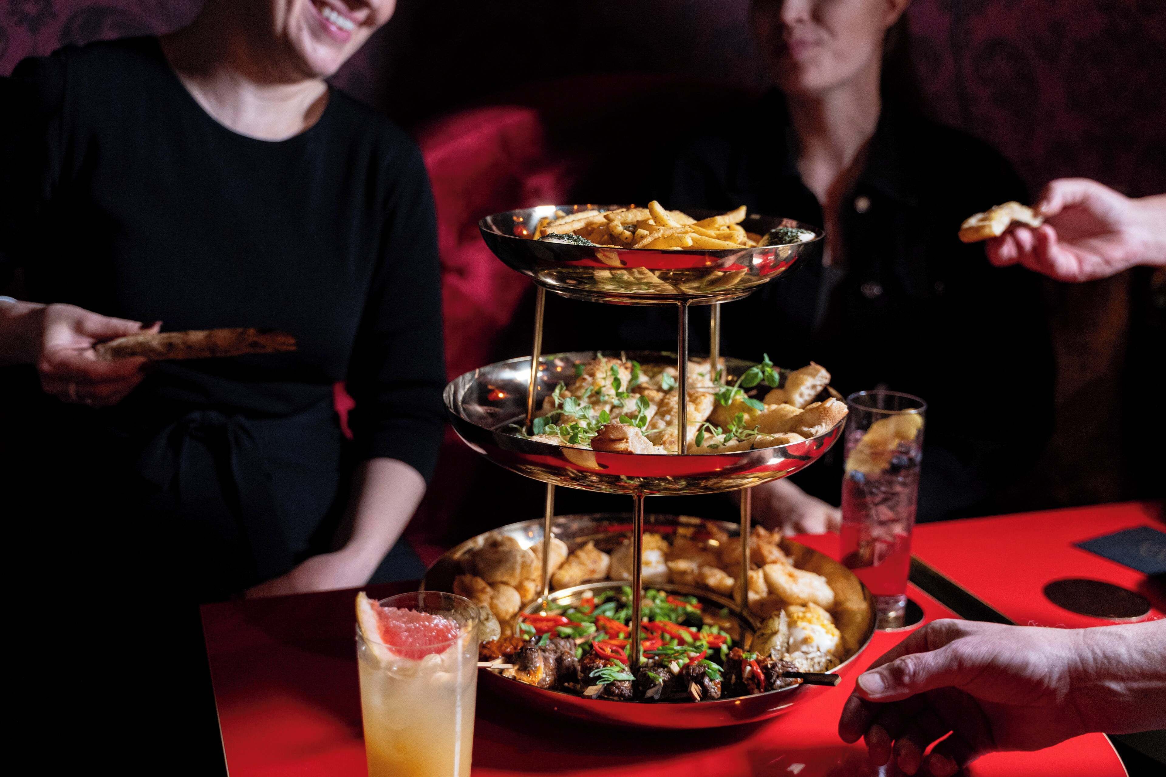 A multi-tiered serving tray with various food items and a drink on a red table.
