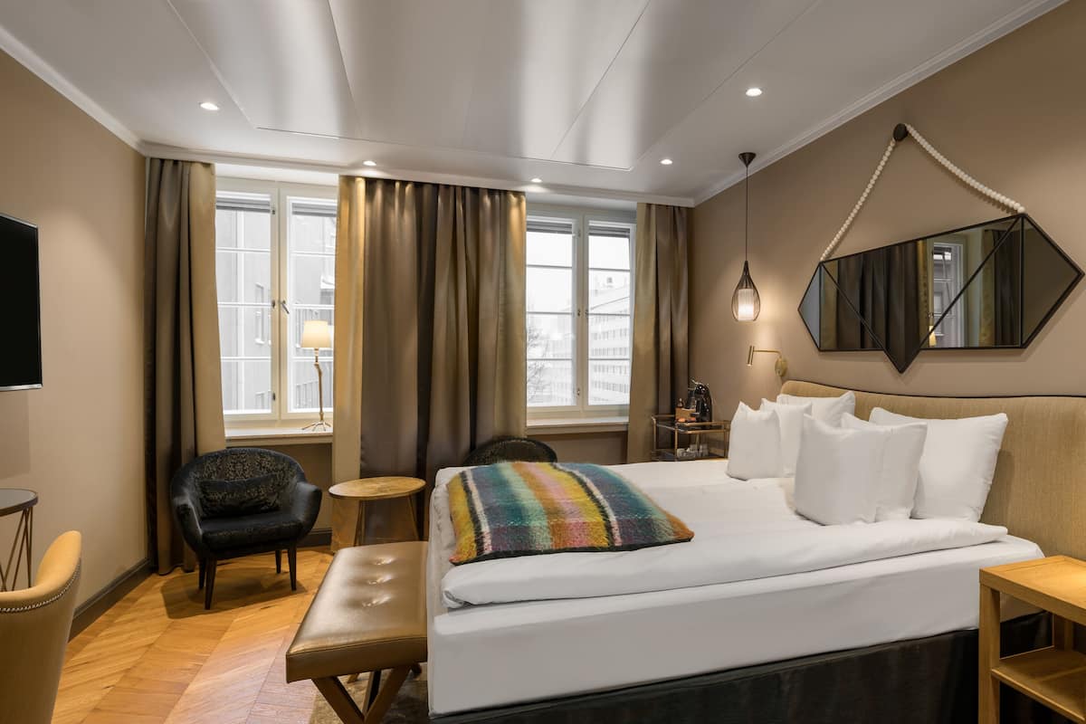 Une chambre d’hôtel avec un lit, une chaise, un bureau, une lampe et une grande fenêtre avec des rideaux.