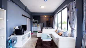 A modern living room with a white sofa, a coffee table, a TV, and a fan. There is a kitchen area in the background with a microwave and cabinets. - Mango Home 102 (Hua Hin)