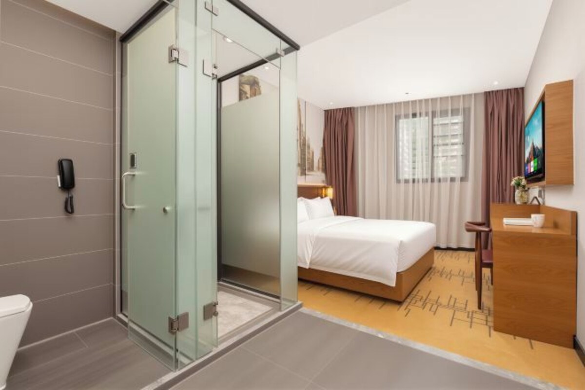 Une chambre d’hôtel dotée d’une douche en verre, d’un lit avec du linge de maison blanc, d’un bureau en bois et d’un téléviseur fixé au mur.