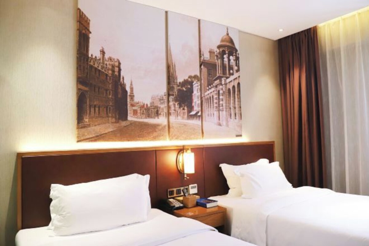 Une chambre d’hôtel avec deux lits, une table de chevet avec une lampe et un grand tableau encadré représentant une vue sur la ville, accroché au mur.