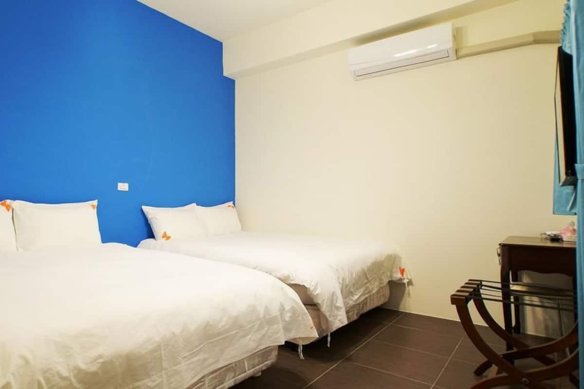 Een hotelkamer met twee bedden, een blauwe accentmuur, een klein tafeltje en een airconditioner.