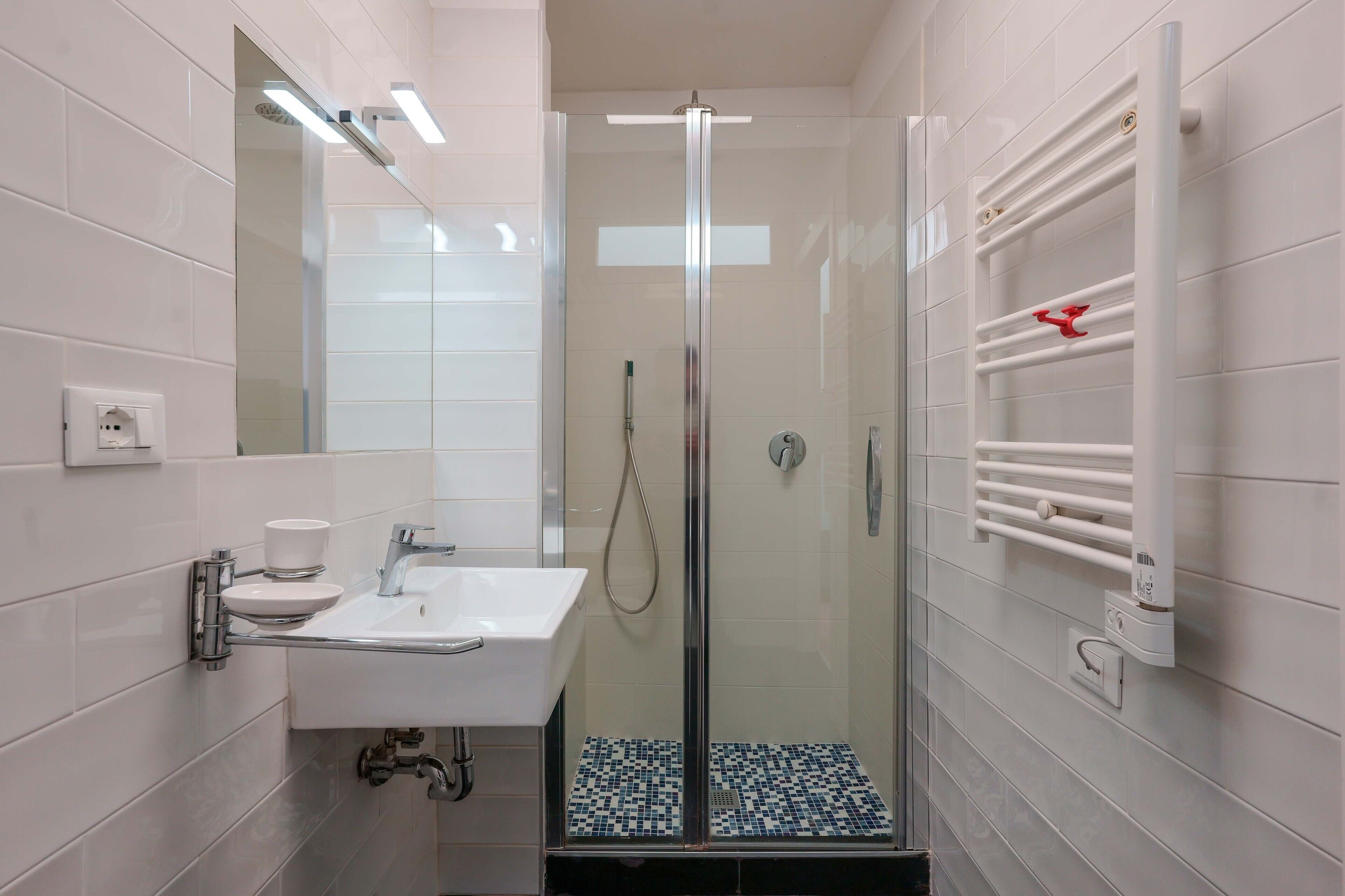 Un baño moderno con ducha de mampara de vidrio, un lavamanos blanco y un perchero con una toalla roja.