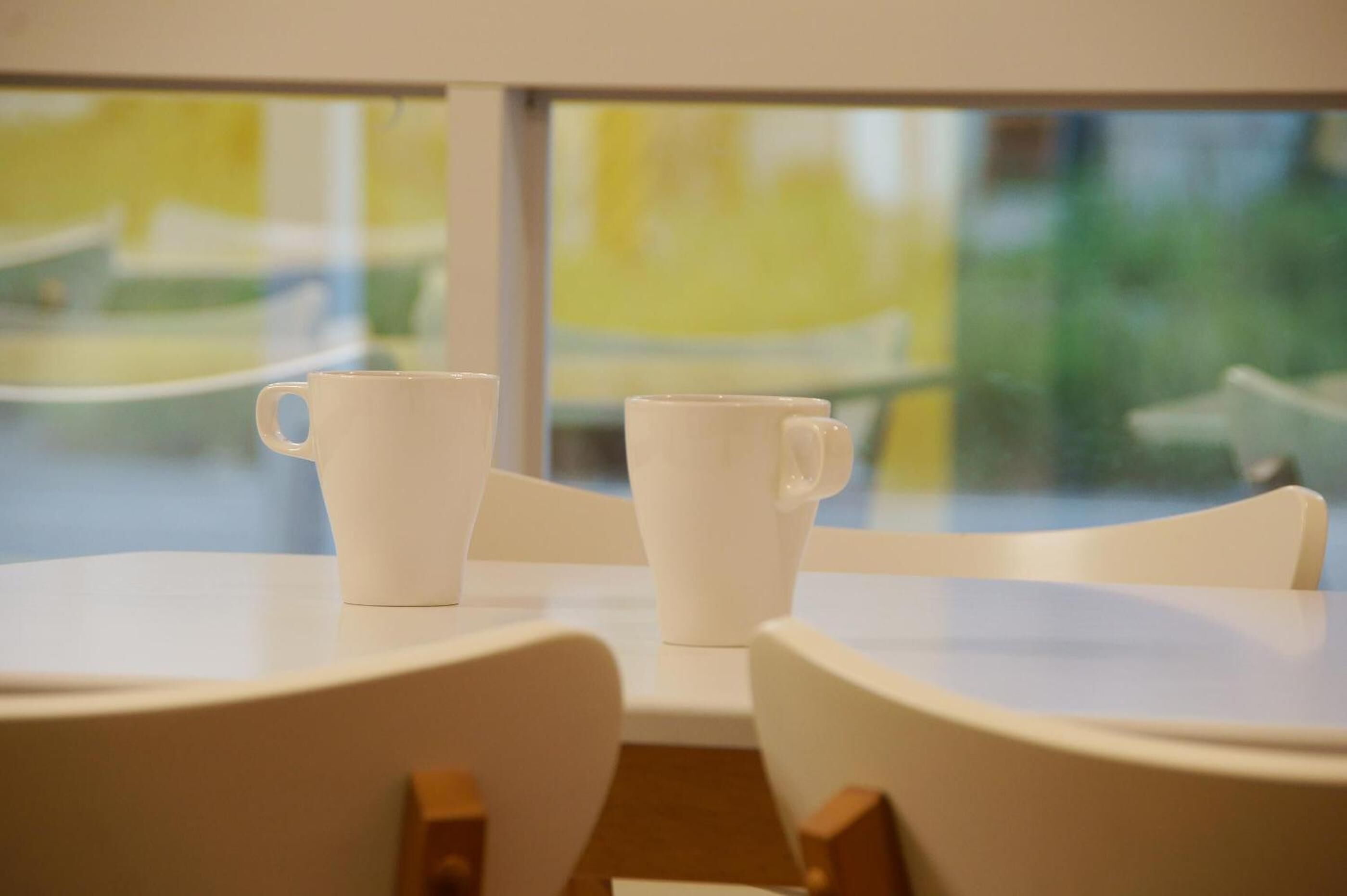 A table with two white cups and chairs in a room with a view through a window.