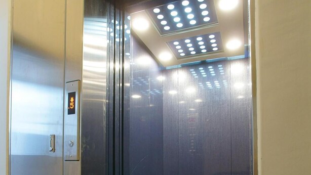 An elevator with a metallic interior, a control panel, and a ceiling with recessed lighting.