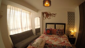 Un dormitorio con cama, sofá, mesita de noche con lámpara, espejo y cabecero decorativo en la pared.