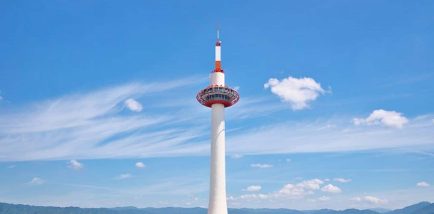Une haute tour blanche, surmontée d’un dôme rouge et blanc, entourée d’un ciel bleu clair et de montagnes lointaines.