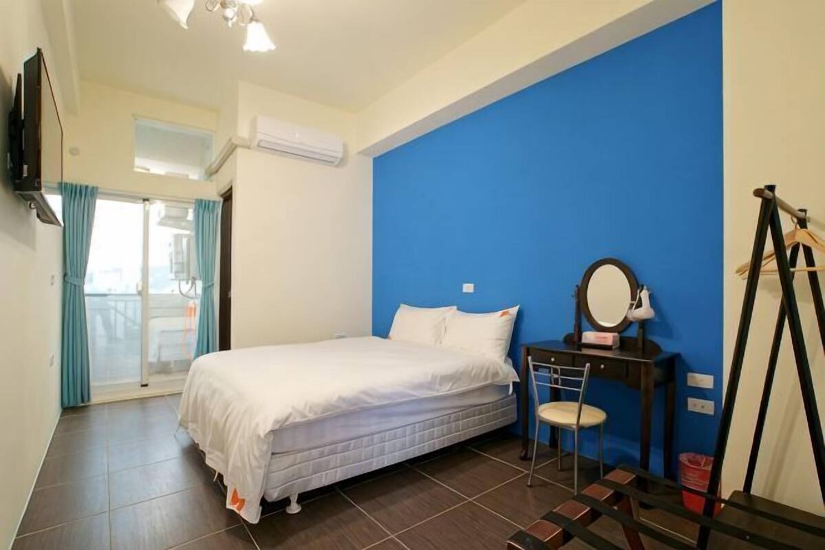 Een slaapkamer met een bed, een bureau met spiegel, een stoel, een raam met gordijnen en een deur die leidt naar een balkon.