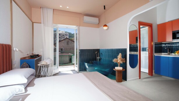 Une chambre d’hôtel moderne dotée d’un grand lit, d’une petite table avec une chaise, d’une salle de bain avec une baignoire en verre et d’un coin cuisine.
