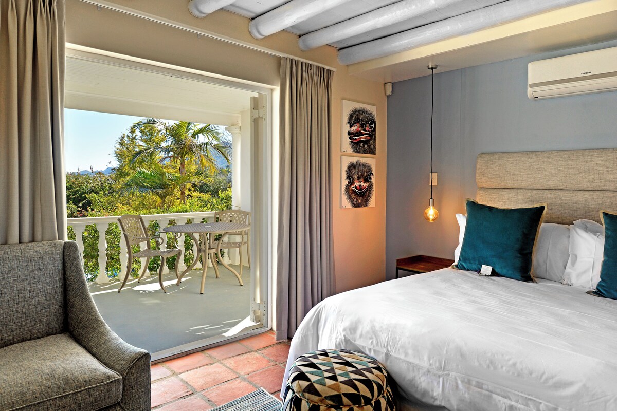 Habitación de hotel con balcón, cama con ropa blanca y dos máscaras tribales colgadas en la pared.