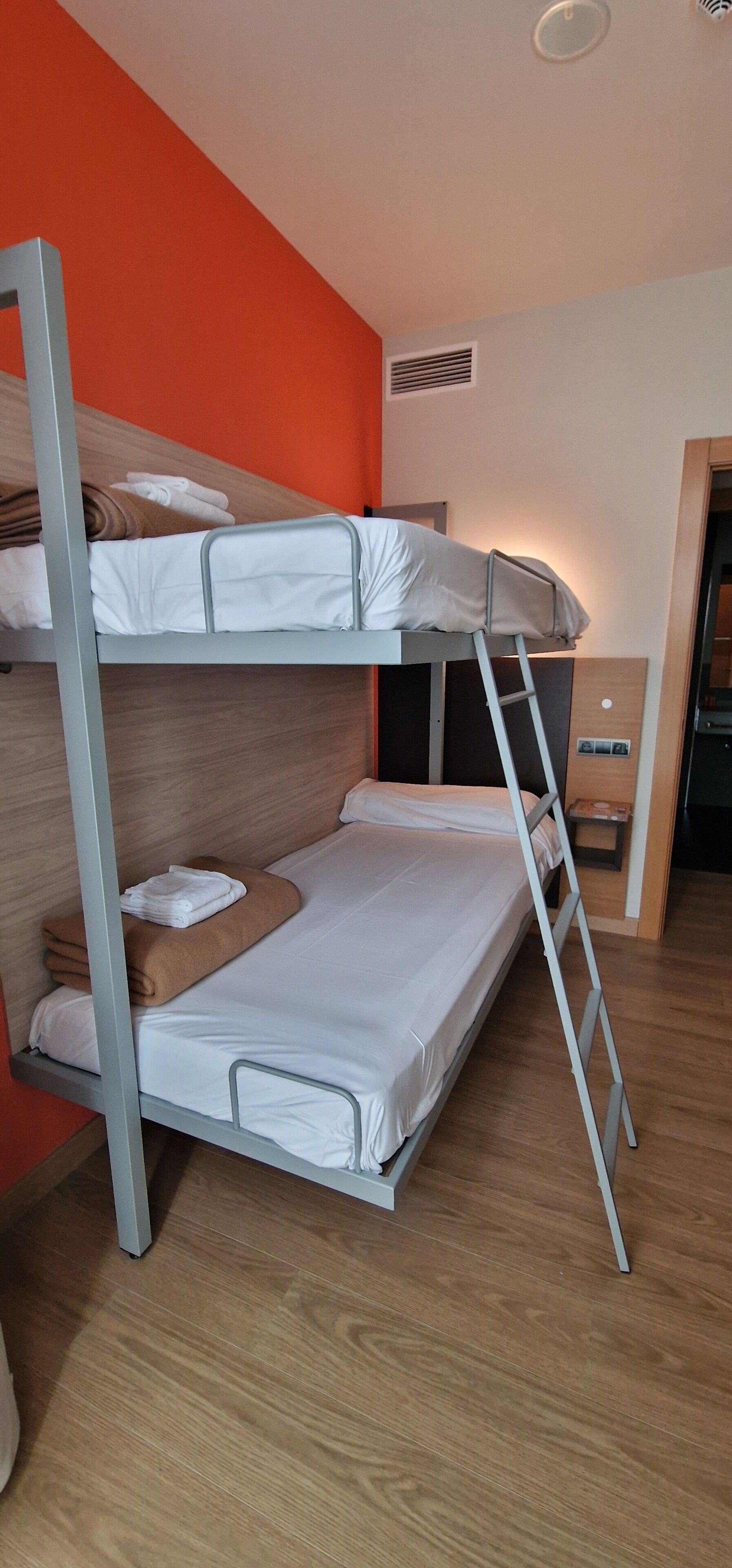 A bunk bed room with a wooden floor, a red wall, and a small table with a lamp.