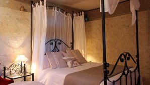 Un lit à baldaquin à quatre poteaux, agrémenté de draps blancs et de coussins décoratifs, une table de chevet avec une lampe et un mur en pierre rustique.