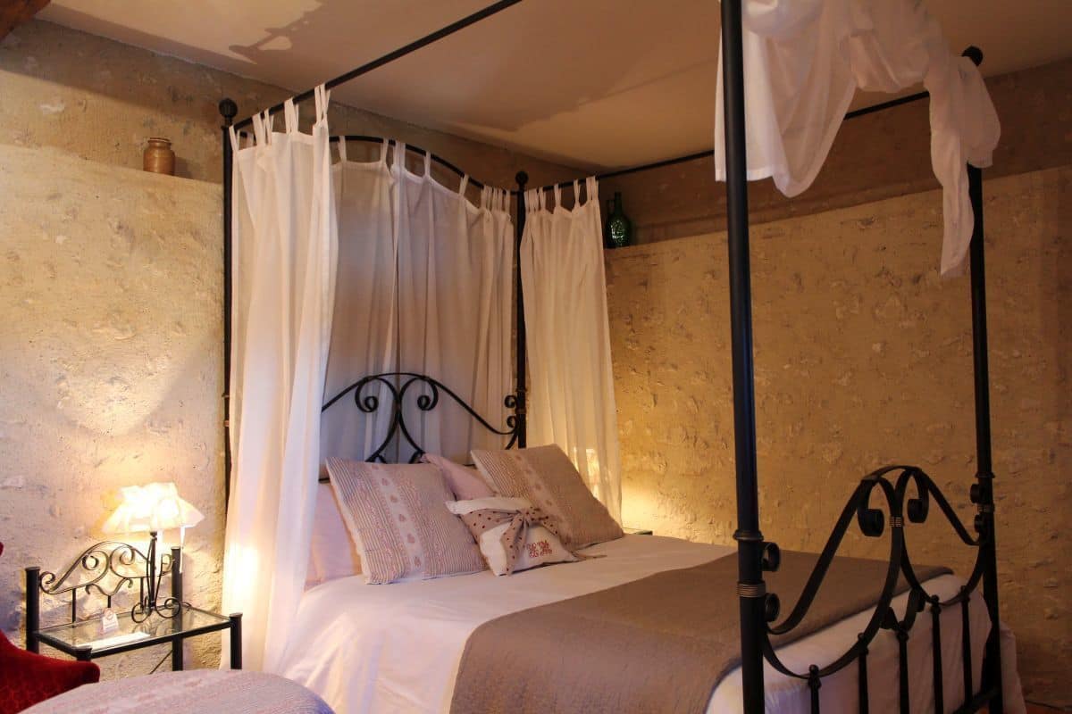 Un lit à baldaquin à quatre poteaux, agrémenté de draps blancs et de coussins décoratifs, une table de chevet avec une lampe et un mur en pierre rustique.