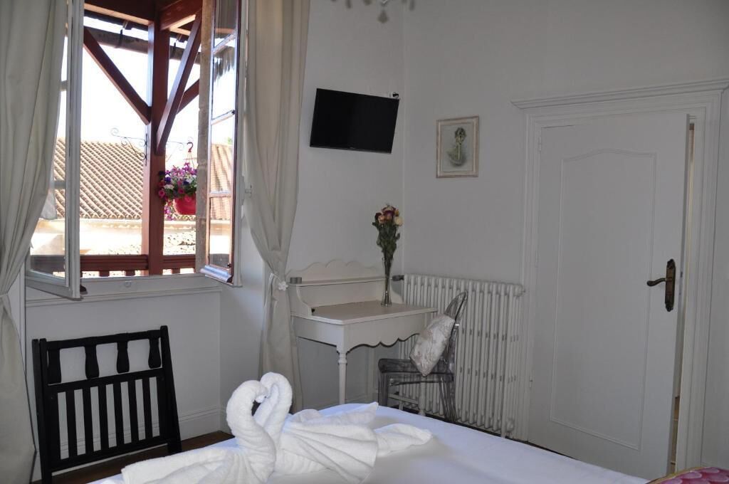 A hotel room with a bed, a small white table, a radiator, a TV on the wall, and a window with curtains.