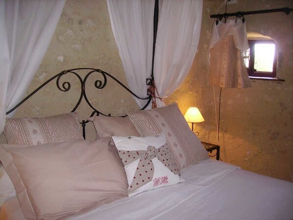 Una cama con estructura metálica, ropa de cama blanca y almohadas estampadas. Se ve una ventana con cortina y una lámpara en la mesita de noche.